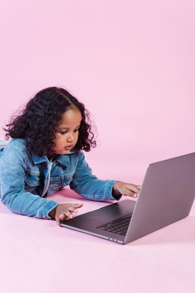 Young girl in denim jacket using a laptop against a pink backdrop.