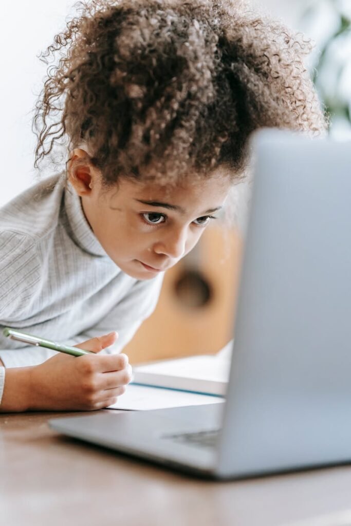 Pensive African American girl with curly hair in casual clothes sitting at table and taking notes while browsing netbook at home