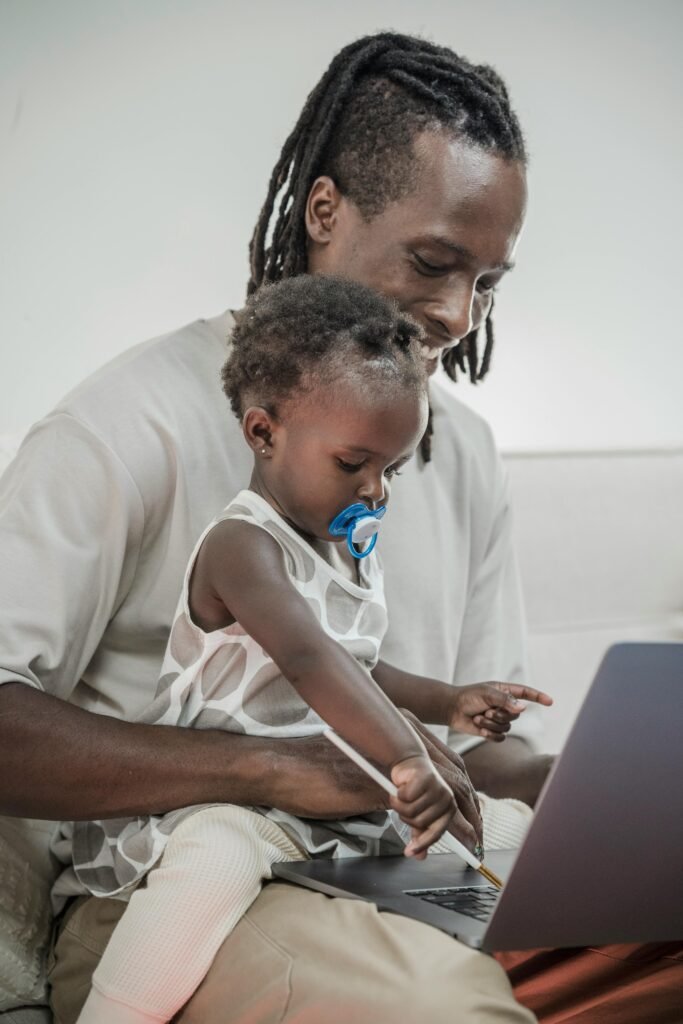 A father with dreadlocks and a baby bonding indoors over a laptop, showcasing modern parenting.